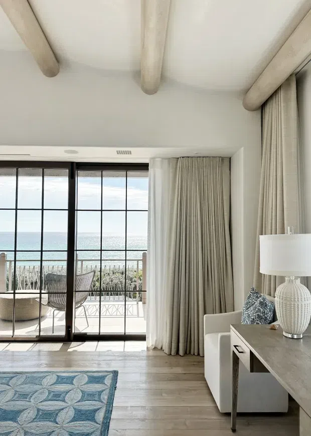 Bright coastal living room with floor-to-ceiling black-framed glazed doors opening to a balcony overlooking the ocean. The room features pale timber flooring, white walls, light exposed rounded ceiling beams, full-height beige curtains, a modern console desk, upholstered chair, table lamp, and a blue patterned rug. Outdoor seating and coastal water views are visible beyond the glazing.