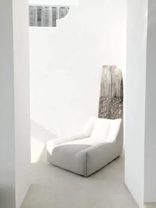 A bright, minimalist white interior viewed through a doorway, featuring a sculptural upholstered lounge chair positioned against a white wall. A tall woven decorative object stands behind the chair, while a small high-level louvred opening adds architectural detail. The space has pale smooth flooring, crisp white walls, strong natural light, and a sparse, serene composition.