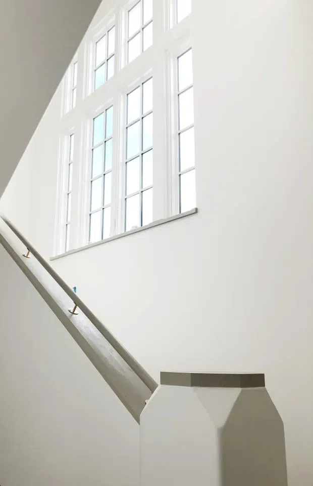A bright, minimalist stairway or stair landing with white walls and tall multi-pane windows. The diagonal staircase has a simple pale railing with small metal brackets, while the high glazing floods the space with natural light. The composition emphasizes vertical window proportions, clean architectural surfaces, and a restrained monochromatic palette.
