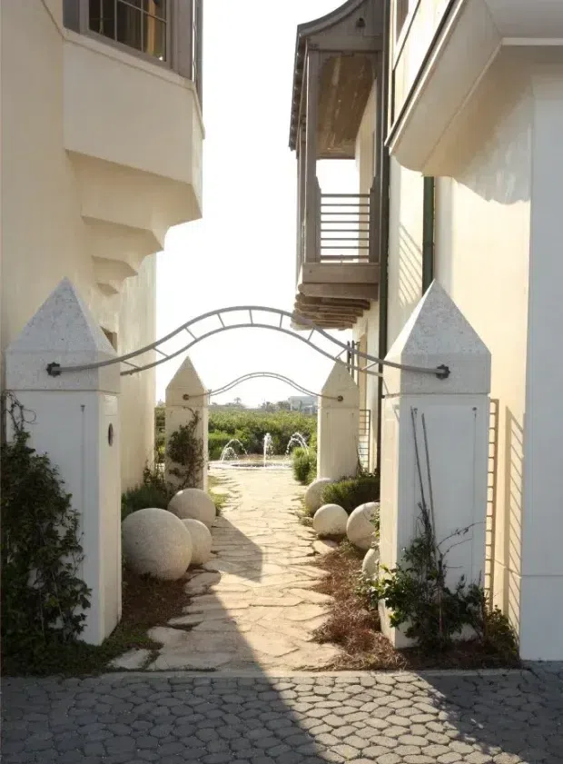 A narrow landscaped passage between white rendered buildings, entered through a decorative gateway with pointed masonry pillars and curved metal gates. The irregular stone path leads toward a small fountain and garden beyond, with spherical stone ornaments and climbing plants along the edges. Upper-level balconies and projecting architectural forms frame the passage, creating a bright Mediterranean-inspired courtyard approach.