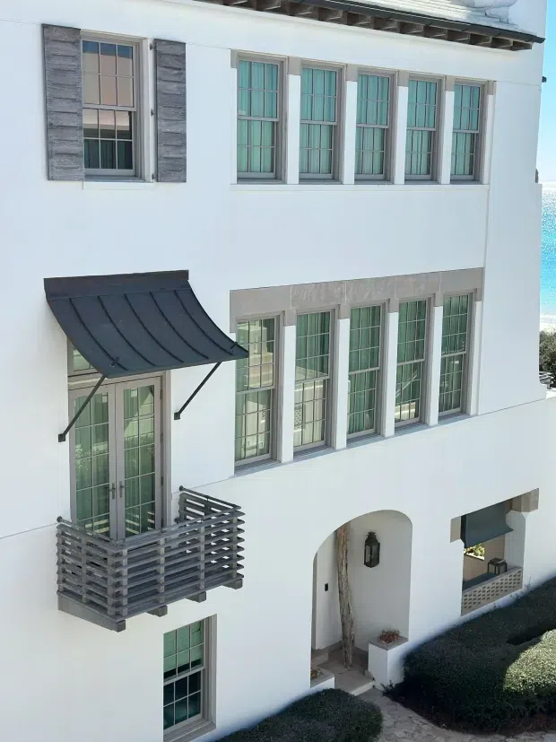 Exterior view of a white multi-storey residence with repeated tall divided-light windows, grey stone or concrete window surrounds and shutters, a dark curved metal awning, and a small projecting balcony with horizontal grey slatted railing. An arched recessed entry and landscaped hedges are visible at ground level. Bright coastal water appears at the far right edge.