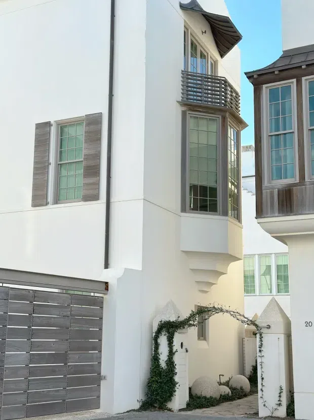 Narrow streetscape view of a tall, white rendered residential facade with a projecting bay window, small balcony with horizontal metal balustrade, decorative curved roof awning, and grey timber-look shutters. The ground level includes a large horizontal-panel garage door and an arched pedestrian entry framed by pointed masonry gate piers and climbing greenery. An adjacent dwelling with dark weathered timber cladding and tall multi-pane windows is visible to the right.