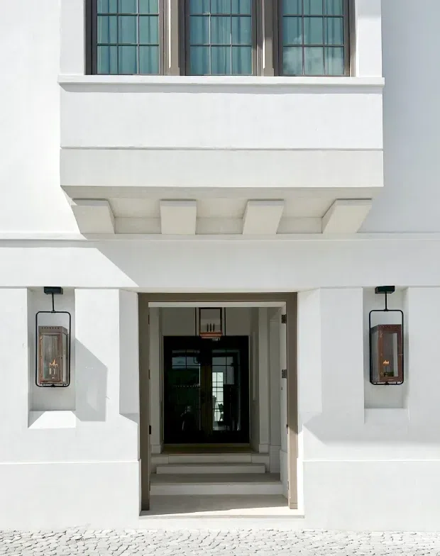Front entrance of a refined white rendered building, featuring a recessed doorway with stepped threshold, symmetrical wall-mounted lanterns, a projecting upper-level bay with decorative corbels, and tall multi-pane windows in dark frames. The foreground is paved with light cobblestones.