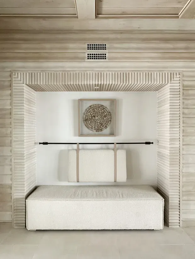 A softly lit, highly symmetrical built-in alcove with a deep upholstered bench or daybed, cream boucle-style upholstery, a slim black hanging rail, and a folded textile suspended behind it. The alcove is framed by elaborate pale ribbed and fluted trim, with whitewashed horizontal timber wall panelling and a panelled ceiling above. A small neutral-toned circular artwork adds a subtle decorative focal point. The overall design is warm, restrained, and textural.
