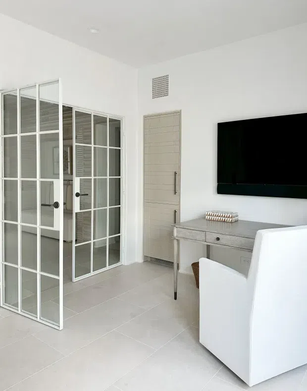 Bright, compact contemporary interior featuring white tiled flooring, white walls and ceiling, a pale grey console desk with drawers, a wall-mounted television, and a white upholstered armchair. A glazed, black-framed grid door opens to an adjacent space, while a light textured internal door and small wall vent add architectural detail. The room has a clean, restrained and highly light-toned appearance.