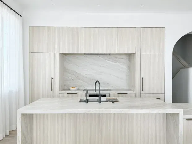 Bright, minimalist kitchen featuring a large central island with an integrated sink and dark arched gooseneck tap. The island and full-height cabinetry are finished in pale vertically fluted surfaces with a light veined stone benchtop. Matching stone is used as the recessed splashback behind the cooktop, framed by extensive handleless and slim-handle storage. Sheer curtains line the left side, while an arched opening is visible to the right.