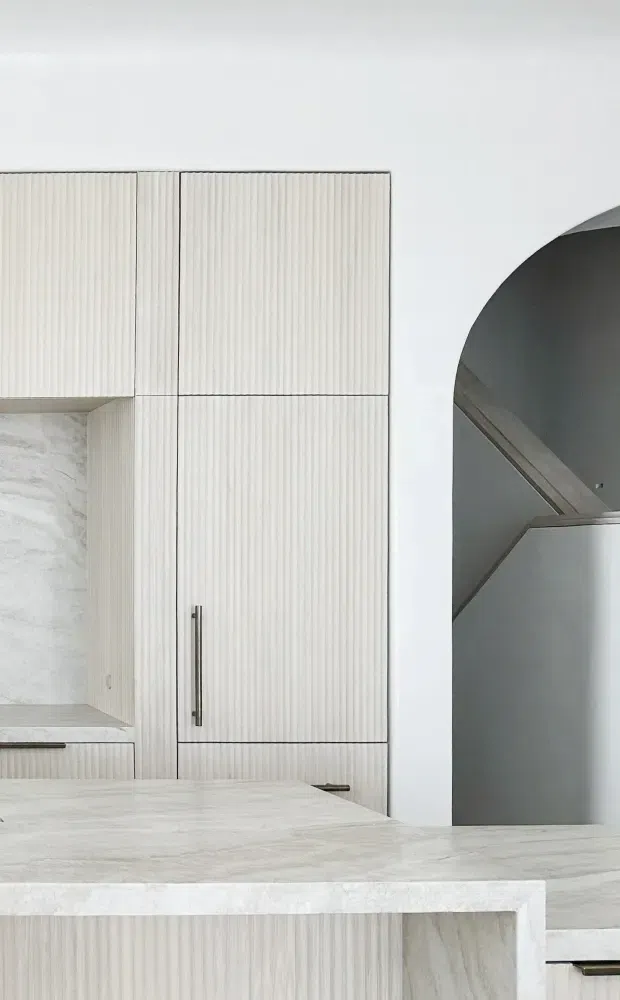 Close-up architectural interior view of a refined, minimalist kitchen. Pale veined stone surfaces form the benchtop, splashback, and island, while tall fluted light-toned cabinetry with slim dark metal handles provides built-in storage. A large arched opening reveals a dark grey stairway or adjoining circulation space. The composition is monochromatic, clean-lined, and strongly texture-focused.