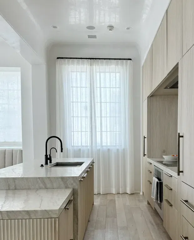 Bright, narrow contemporary kitchen with a marble-look waterfall island and integrated sink, pale vertically fluted cabinetry, concealed appliances, and full-height sheer curtains over a large gridded window. The space has a clean, light-toned palette and a polished, refined finish.