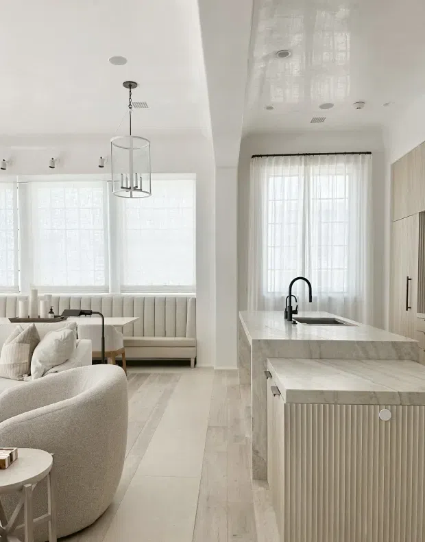 Bright open-plan interior showing a contemporary kitchen with a large pale stone island and fluted light cabinetry, opening directly into a refined living area with curved upholstered seating, built-in banquette, sheer window coverings, and a linear pendant light. The space has white walls and ceilings, pale timber flooring, abundant filtered daylight, and a soft neutral palette.