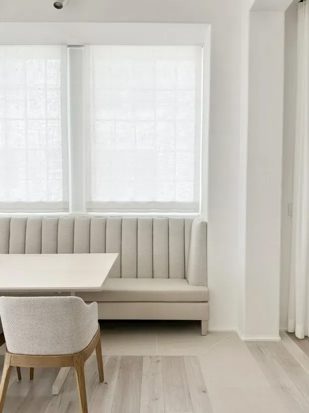 Bright, softly styled dining nook with a built-in upholstered banquette beneath large windows dressed in sheer white roller shades. A pale rectangular dining table and textured upholstered wooden chair sit on light timber-look flooring. White walls, deep window reveals, simple trim, and recessed ceiling lighting create a clean, warm minimalist interior.