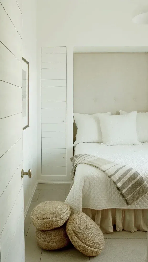A narrow, bright bedroom with a soft neutral palette. The room features a white upholstered bed with layered white pillows and quilted bedding, a taupe upholstered headboard, a striped grey-beige throw, white horizontal wall panelling, a tall louvred or panelled cupboard door, and pale tiled flooring. Woven round ottomans sit at the foot of the bed, creating a calm, textural coastal-inspired interior.