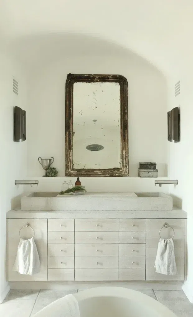 A bright, minimalist bathroom vanity with a large double-basin stone or composite countertop, extensive pale built-in cabinetry, wall-mounted taps, and a freestanding bath visible in the foreground. A weathered dark timber-framed mirror, sculptural wall sconces, symmetrical towel rings, and restrained decorative accessories add a rustic traditional contrast to the clean white room.