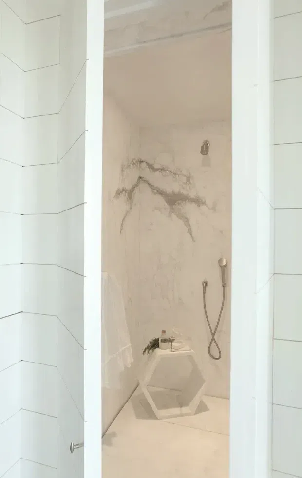 A narrow, minimalist shower room viewed through an open white door. The shower enclosure is lined in pale veined marble or stone slabs, with a wall-mounted overhead shower and handheld shower rail. A small geometric hexagonal stool or side table sits inside, and the adjacent white door surfaces feature subtle angular panel lines.