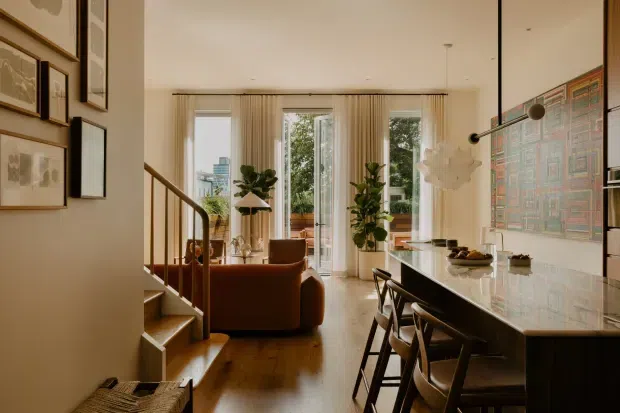 Warm, refined open-plan interior combining a kitchen, dining counter and living area. The space features dark wood flooring, a large pale stone kitchen island with integrated seating, timber stair detailing, full-height glazed doors with sheer curtains, framed artwork, indoor plants and sculptural pendant lighting. A living area is visible beyond the island, with a garden or terrace seen through the glazing.