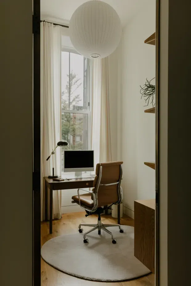 A compact, softly lit home office viewed through a doorway, featuring a dark wood desk with a computer and desk lamp, a brown leather office chair, tall windows with sheer curtains, built-in wood shelving, a large spherical pendant light, warm timber flooring, and a round neutral rug.