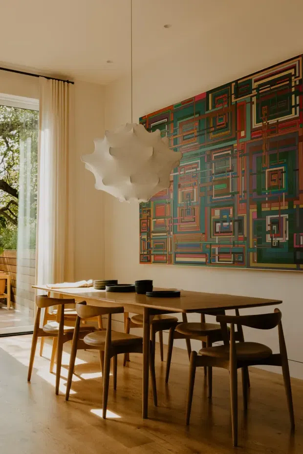 A warm, sunlit dining room with a long solid-wood dining table and matching wooden chairs. A large colourful geometric abstract artwork dominates the wall, while a sculptural white pendant light hangs above the table. Full-height curtains and a large window bring in views of trees, and the room has pale walls and timber flooring.