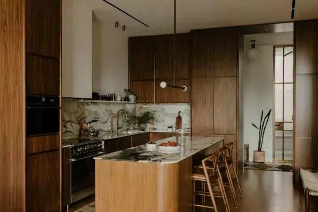 A refined contemporary kitchen with dark timber cabinetry, a veined marble backsplash and island benchtop, integrated appliances, and warm timber flooring. The island includes a curved timber-clad base and seating for several people, while a glazed doorway leads to an adjacent area.