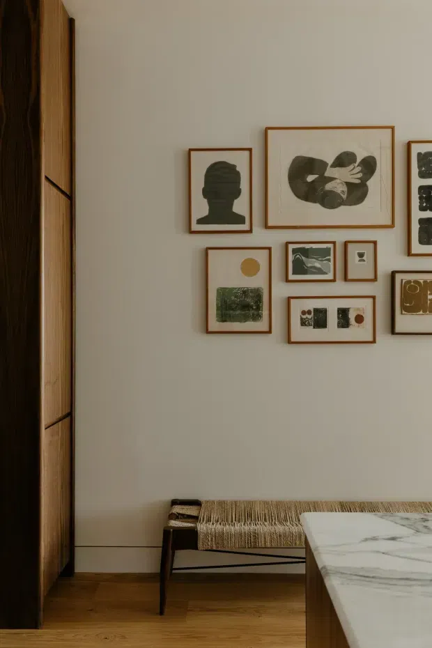 A warm, refined interior vignette featuring a pale neutral wall arranged with a gallery of framed abstract and graphic artworks. A dark timber and light timber built-in joinery element occupies the left side, while a low woven bench sits against the wall. In the foreground is the corner of a substantial marble-topped timber table or island. Timber flooring and warm, subdued lighting create a calm, curated atmosphere.