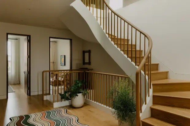 Interior view of a refined residential stair hall with a sweeping curved staircase, warm timber stair treads and handrail, slender vertical balusters, and a matching upper-level balustrade. Pale walls, timber flooring, framed artwork, potted greenery, and a curved patterned rug create a warm, elegant transitional setting. A doorway and bright adjacent room are visible to the left.