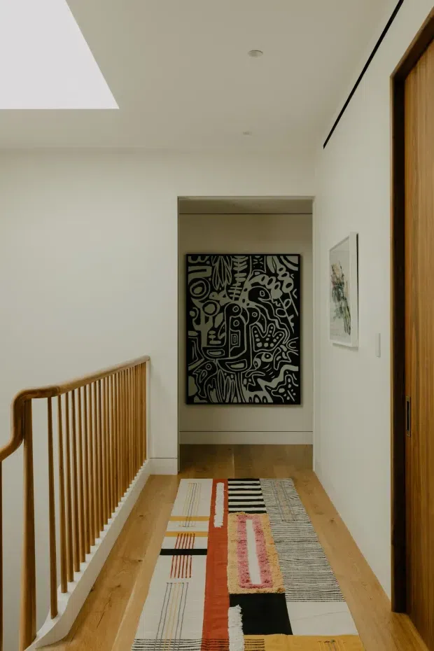 A bright, narrow upper-level hallway or landing with pale walls, warm timber flooring, a timber balustrade overlooking the stairwell, and a full-height timber door at right. A colourful geometric runner extends toward a recessed doorway or alcove with bold black-and-white artwork. Minimal artwork and discreet recessed ceiling lights create a clean contemporary interior.