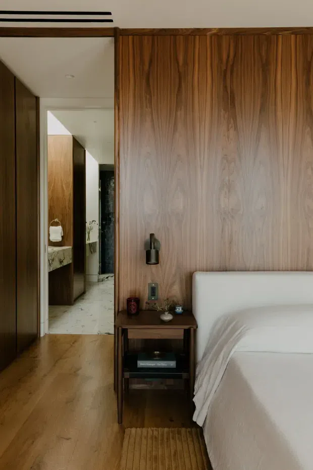 Warm, refined bedroom interior featuring a bed with a white upholstered headboard and white bedding, a dark timber bedside table, and extensive vertical-grain timber wall panelling. The room has matching timber flooring, a small bedside wall light, a textured neutral rug, and a full-height mirror or glazed opening reflecting a hallway and adjacent bathroom vanity area. The overall look is calm and contemporary with strong natural wood detailing.