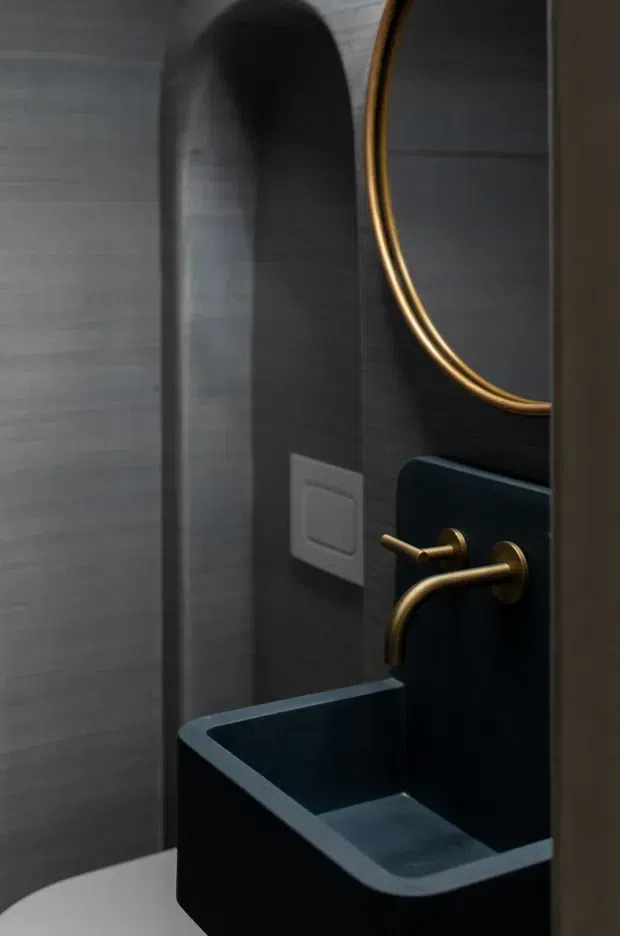 A compact, dark-toned bathroom viewed through a doorway, featuring a deep navy or black integrated wall-mounted basin with rounded edges, brushed brass wall-mounted tapware, a large round brass-framed mirror, dark grey tiled walls, and a white light switch. The space has a moody, refined contemporary feel with a narrow arched wall recess or doorway detail.