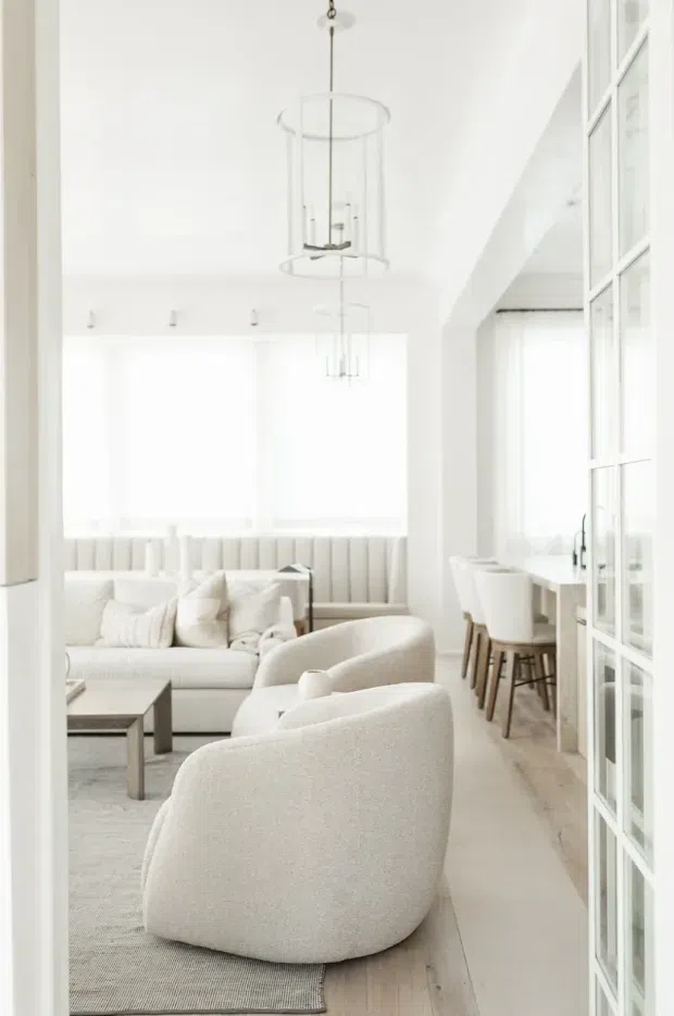 Bright, predominantly white open-plan living and dining space with a soft contemporary interior. The foreground contains sculptural upholstered lounge chairs and a pale sofa around a simple coffee table, while a dining counter with stools sits beside tall multi-pane glazing or framed internal doors. Sheer window coverings, fluted wall detailing, pale timber flooring, and delicate cylindrical pendant lights create an airy, refined atmosphere.