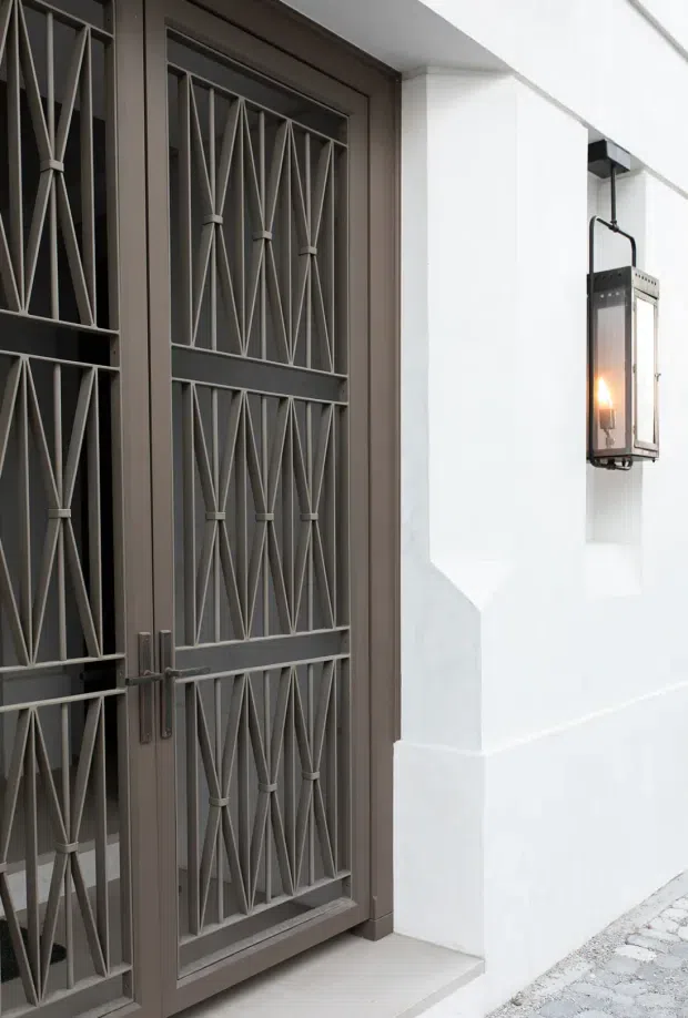 Close-up exterior view of a contemporary residential entrance. A dark grey metal security door with repeated geometric diamond-pattern grillework is set into a white rendered wall, with a matching rectangular wall-mounted lantern and a light tiled threshold. A small section of pale cobblestone paving is visible at the base.