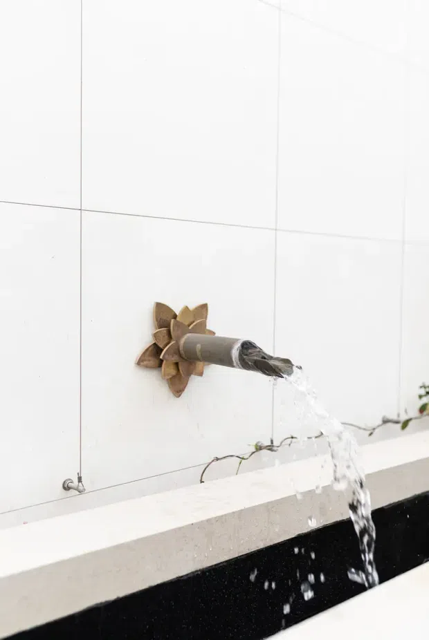 Close-up of a sculptural wall-mounted water spout shaped like a flower, pouring into a long dark basin or water feature. The surrounding wall is clad in large white tiles with fine grout lines, and a pale stone or concrete ledge runs along the basin edge. A small vine is visible near the water feature.