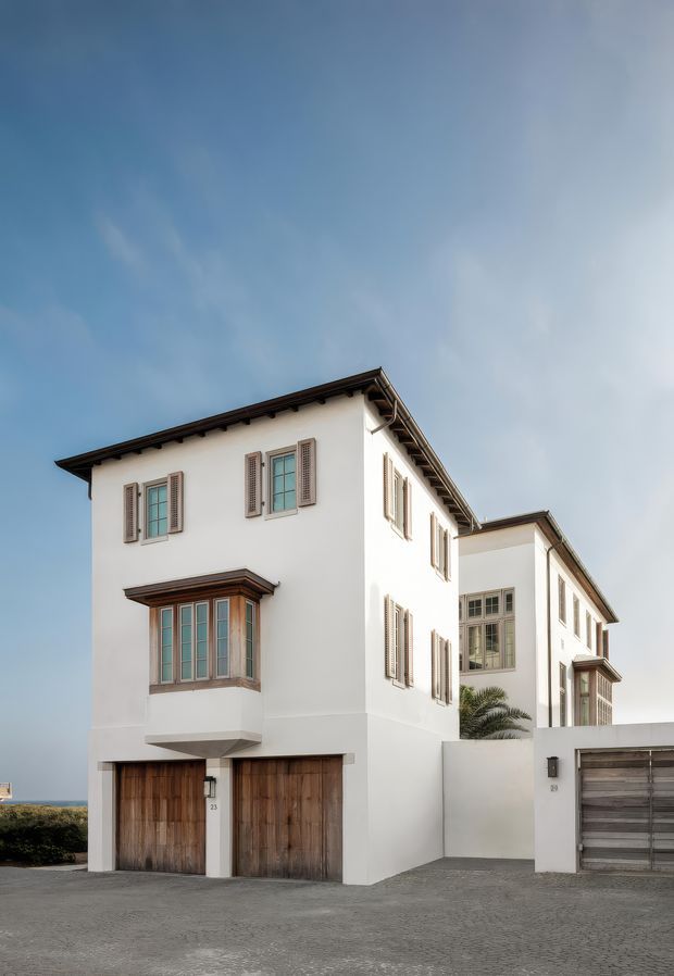 Exterior view of a multi-storey white stucco residence with dark pitched roof eaves, timber-framed windows, decorative exterior shutters, projecting bay window, and multiple timber garage doors. The home sits along a paved driveway, with a glimpse of coastal water visible in the distance.