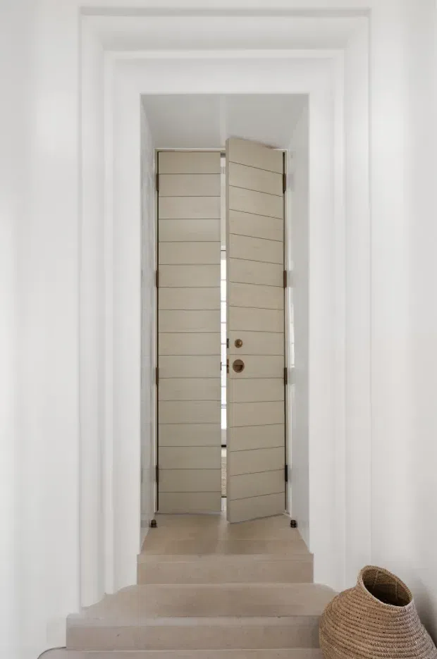 A minimalist interior view of a tall white-painted entry passage with broad stepped flooring leading to a partially open, contemporary double front door. The doors feature pale horizontal timber panels, slim dark hinges, and bronze-toned hardware. White walls and trim create a bright, restrained composition, with a woven basket placed at the lower right.