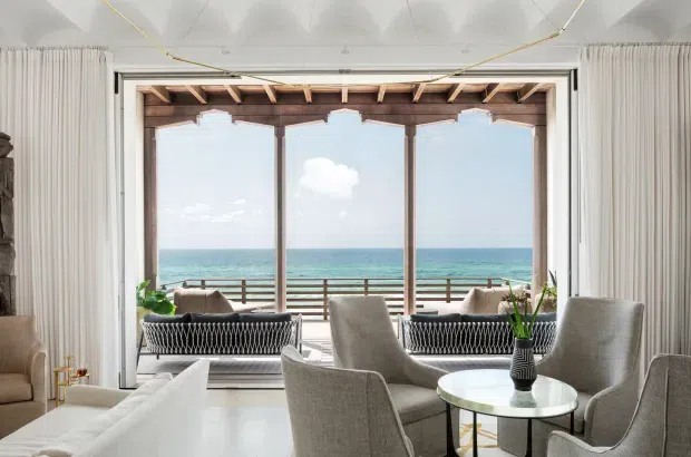 Bright coastal living room with a refined seating arrangement facing expansive ocean views through large folding or sliding glazed doors. The room features light neutral upholstered armchairs and sofa seating, a round glass coffee table, white curtains, pale flooring, and a distinctive exposed timber beam and rafter structure above the opening. A balcony or terrace with timber railings is visible beyond the glazing.
