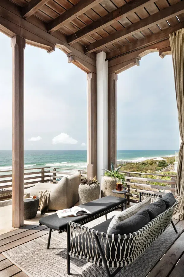 A bright coastal outdoor lounge or enclosed terrace overlooking the ocean. The space features exposed timber roof beams and decking, substantial timber posts, white structural columns, open glazed or screened sides, a woven rope-framed sofa with dark cushions, a black coffee table, textured neutral rug, planters, and full-height curtains. The ocean and beach are visible beyond the balustrade.