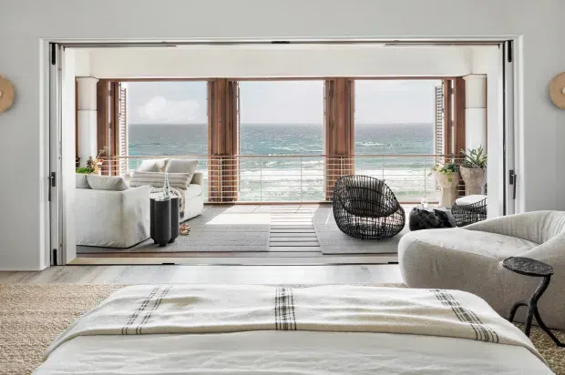 A bright coastal bedroom opening onto a sea-facing sunroom or balcony through wide folding glass doors. The room features a large upholstered bed in the foreground, neutral bedding and seating, timber-framed glazing, and a relaxed sitting area with ocean views beyond.