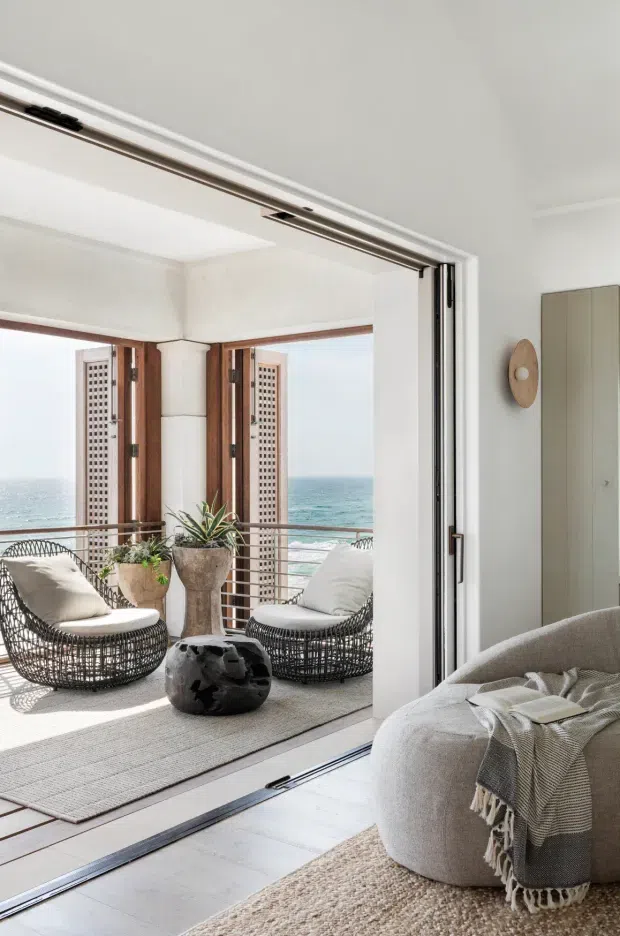 Bright coastal living room opening onto a balcony or terrace with ocean views. The room features white walls and ceiling, pale timber flooring, a large upholstered sofa, textured rugs, woven lounge chairs, a sculptural dark coffee table, plants, and wide timber-framed folding or sliding doors. A perforated privacy screen and metal balcony railing are visible outside.