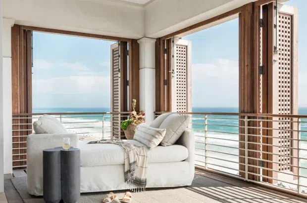 Bright coastal-facing living area with a white upholstered sofa, soft cushions and throw, dark cylindrical side table, and potted arrangement. Large timber-framed corner glazing opens to a balcony with horizontal metal balustrades and expansive ocean views. Decorative perforated privacy screens flank the windows, and strong sunlight creates linear shadows across the carpet.