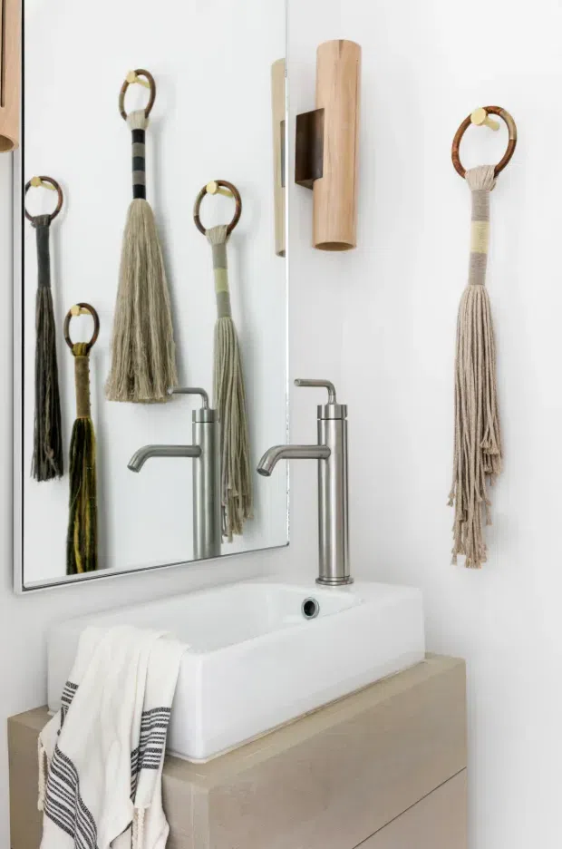 A compact, bright bathroom vanity with a white rectangular vessel sink mounted on a pale wood-toned floating cabinet. Brushed metal tapware sits in front of a large frameless mirror, which reflects the sink and decorative hanging tassels. A timber wall-mounted cylindrical light fixture and multiple neutral-toned textile tassels add handcrafted detail to the white walls.