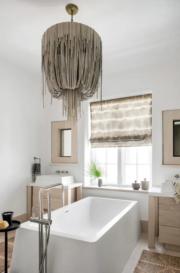 Bright, refined bathroom featuring a freestanding white bathtub beneath a dramatic cascading-strand pendant light. The room includes twin white vessel basins on light timber-toned vanities, chrome tapware, framed mirrors, a large window with a textured Roman blind, and neutral woven rugs and accessories.