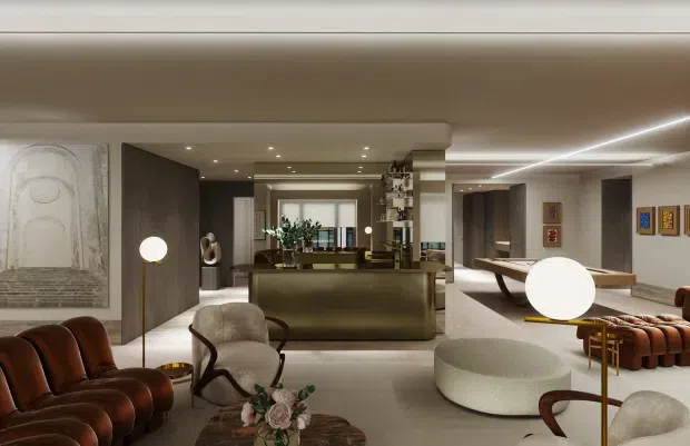 A spacious, refined contemporary living area viewed toward an open bar or kitchen zone. The room features warm neutral finishes, dark timber wall panels, a large central island or counter, recessed ceiling lighting, sculptural upholstered seating, leather lounge chairs, a rounded ottoman, and decorative artwork. A linear illuminated ceiling detail and open shelving add modern character.