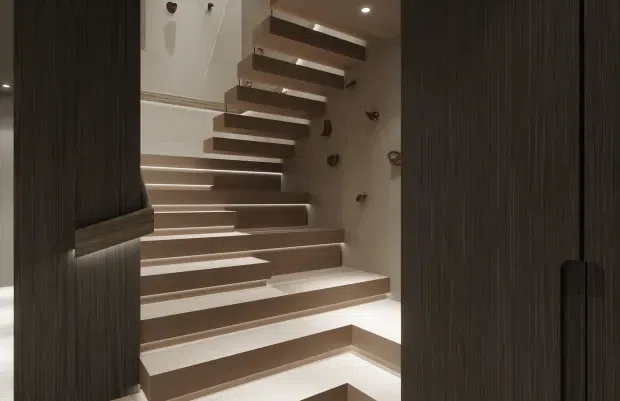 A contemporary stairway viewed through dark timber-textured joinery or doors. The staircase has broad floating or cantilevered steps in contrasting light and dark finishes, integrated step lighting, and a sculptural wall with small decorative protrusions. Recessed ceiling lighting and minimalist detailing create a dramatic, refined interior.
