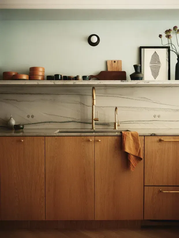 A warm, carefully styled kitchen featuring full-height walnut-toned timber cabinetry with slim brass pulls, a pale veined stone splashback and benchtop, and a brass gooseneck tap with a separate drinking-water tap. An open display shelf holds ceramics, cutting boards, pottery and framed artwork, while a folded rust-coloured tea towel adds a warm accent. The room has a refined mid-century-inspired character with understated contemporary detailing.