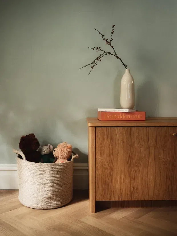 A softly lit interior vignette featuring a low mid-century-style wooden sideboard against a muted sage-green wall. A cream ceramic vase with delicate branches and a red-orange book sit on top, while a woven basket filled with plush toys rests beside the cabinet. Light timber flooring is laid in a herringbone pattern, creating a warm, calm and child-friendly atmosphere.