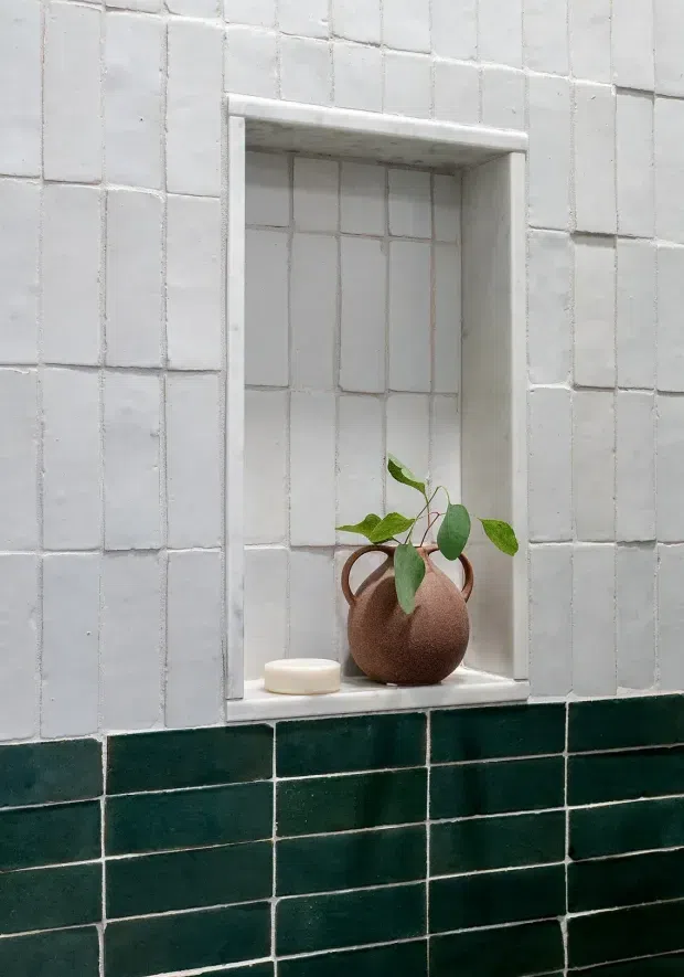 Close-up of a recessed wall niche lined with white rectangular tiles, containing a rounded brown ceramic vase with green foliage and a small white soap dish. The surrounding wall transitions from white tiles above to dark forest-green tiles below, with light grout lines. The image is a minimalist architectural detail, likely in a bathroom or wet area.