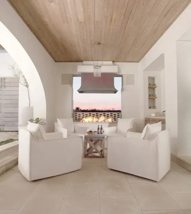 A bright, refined living room arranged around a central fireplace and large rangehood-like feature, with four white upholstered armchairs, a small timber-and-metal coffee table, and built-in shelving. The room has a white plastered interior, pale large-format tiled floor, and a prominent warm timber-lined ceiling. Arched openings connect to an outdoor area, while sunset light is visible beyond the rear opening.