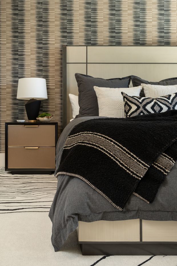 A warmly styled bedroom with a large upholstered or paneled bed featuring dark trim, layered charcoal and cream bedding, and a bold black-and-white geometric throw pillow. A dark timber bedside table with brass-toned drawer pulls supports a sculptural black lamp. The room has a highly textured striped feature wall, neutral carpeting, and a monochrome patterned rug, creating a sophisticated, graphic, high-contrast interior.