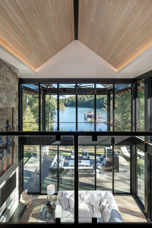 A dramatic double-height living room overlooking a wooded lake through extensive black-framed floor-to-ceiling glazing. The room features a vaulted ceiling clad in light timber boards with concealed linear lighting, a large natural stone feature wall, dark structural framing, and layered living areas visible across the split-level or mezzanine-like composition.