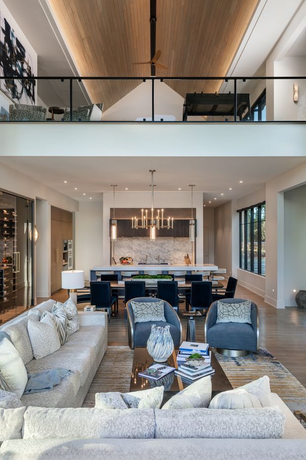 A sophisticated open-plan living and dining area viewed from the seating zone, with a large sectional sofa, blue upholstered lounge chairs, a dining table, and a contemporary kitchen beyond. The room features a dramatic double-height volume with a timber-lined pitched ceiling, upper-level mezzanine, black-framed glass balustrade, large windows, marble-look kitchen splashback, dark timber flooring, and layered pendant and recessed lighting. The palette is neutral and refined with navy-blue accents.