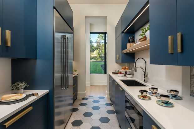 A narrow, contemporary galley kitchen with deep navy-blue cabinetry, brass-toned handles, white benchtops, integrated stainless-steel appliances, open timber shelving, and a white tiled backsplash. A patterned hexagonal floor in white, grey, and blue leads to a tall window overlooking greenery. Under-cabinet lighting and decorative tableware add a refined, inviting feel.