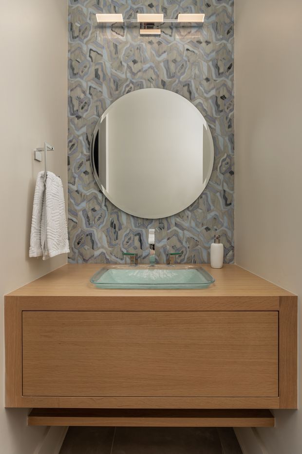 A compact, refined bathroom vanity viewed straight on. The floating timber vanity has a broad natural wood benchtop, an integrated clear glass vessel basin, and wall-mounted tapware. Above it is a large round mirror set against a full-height patterned mosaic tile feature wall, with a horizontal contemporary light fixture. White towels hang on a wall rail at left and a small dispenser sits on the counter.