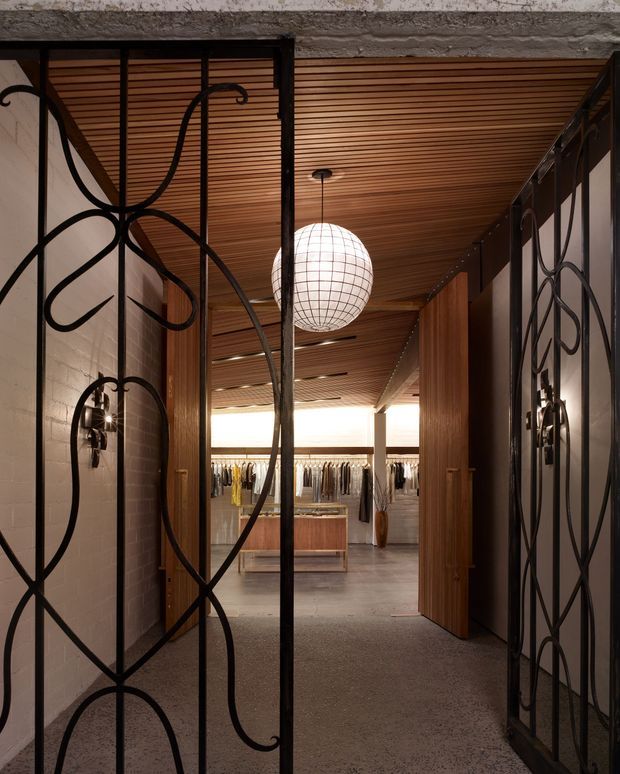 A dramatic interior entrance to a boutique retail clothing space, viewed through ornate black wrought-iron screen doors. The corridor features exposed white brick walls, warm timber slatted ceiling, dark timber doors and panels, concrete or terrazzo flooring, and a large spherical wire pendant light. Clothing racks and a central timber display island are visible in the room beyond.